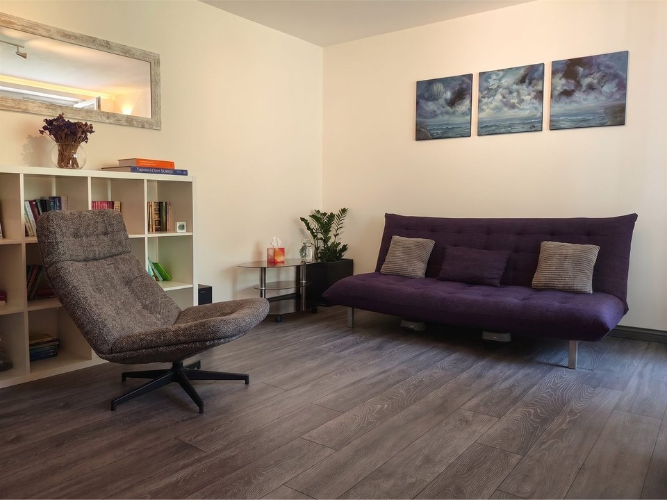Camera de terapie: fotoliu rotativ, canapea, bibliotecă și tripticul agățat pe perete.